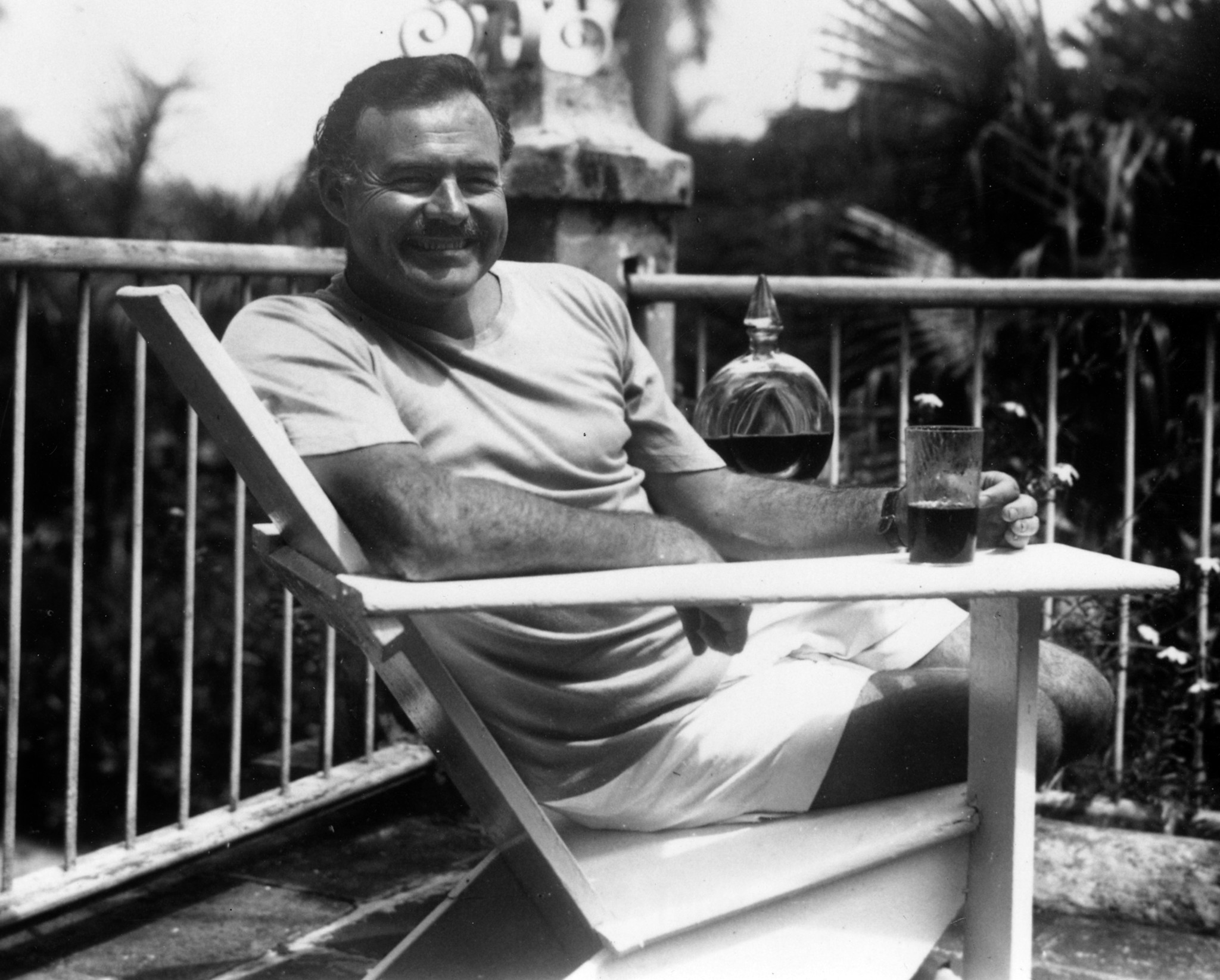 A man with short hair and a mustache is sitting in a chair outdoors, smiling, with a glass and a bottle on a table beside him.