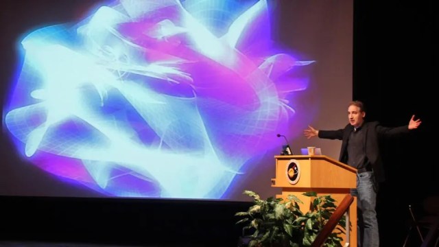 A person is giving a presentation at a podium with a large abstract, colorful light pattern displayed on a screen behind them.