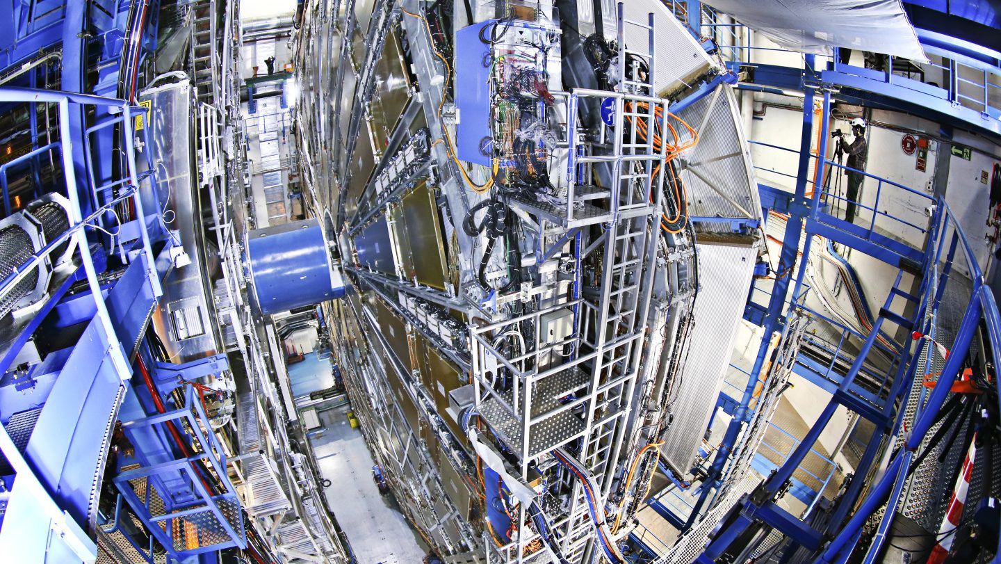 A large, intricate machine with metallic components and blue scaffolding in a laboratory setting. Numerous cables and pipes are connected to the central structure.