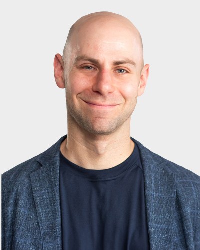 Bald man with a slight smile wearing a blue jacket over a dark shirt, standing against a plain, light-colored background.