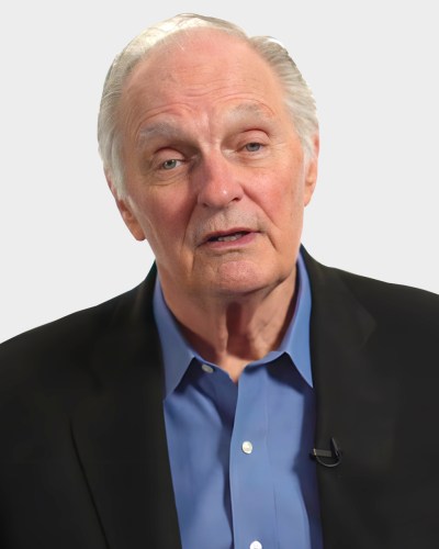 An older man with short gray hair and a receding hairline, wearing a blue shirt and a dark blazer, speaks while facing forward.