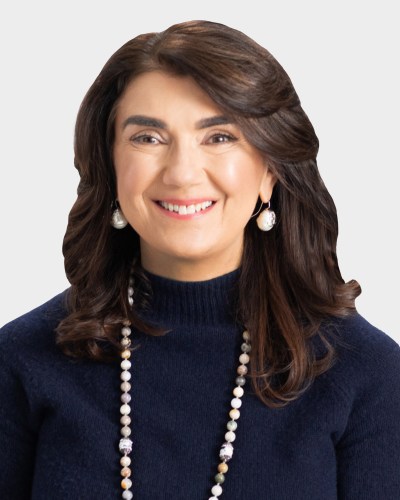 Portrait of a woman with shoulder-length brown hair, wearing a dark sweater, pearl earrings, and a beaded necklace, smiling.