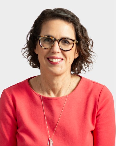 A woman with short, curly hair and glasses is wearing a coral top and a necklace, looking at the camera and smiling.