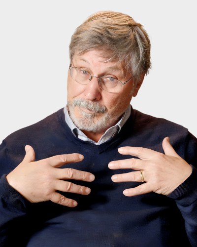 A man with glasses, a beard, and gray hair gestures with both hands while wearing a dark sweater and a shirt, standing against a plain background.