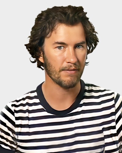 A man with wavy hair and a beard, wearing a black and white striped shirt, looks at the camera against a plain background.