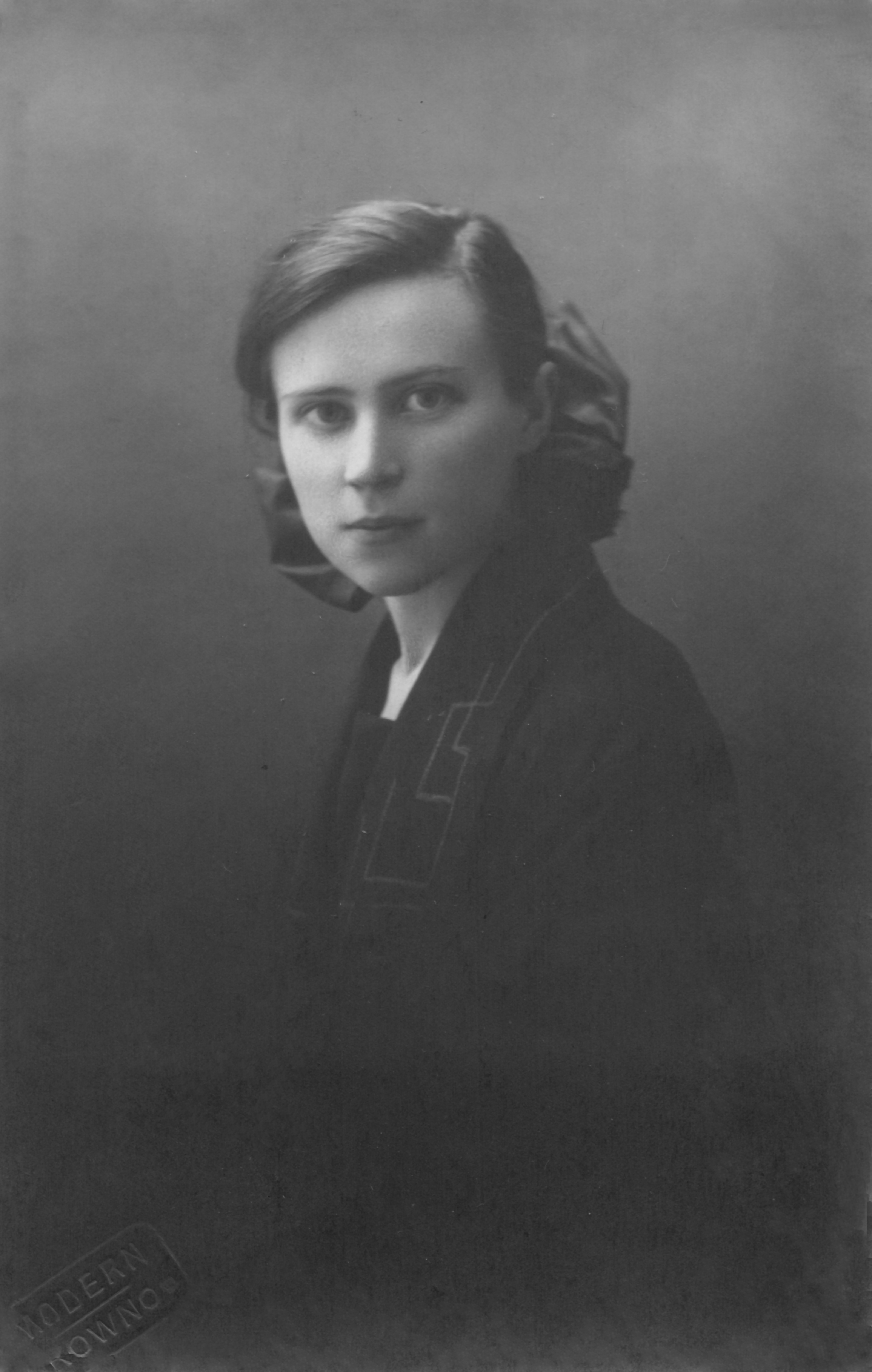 Black and white portrait of a woman with short hair, wearing a dark outfit and a large bow at the back of her head, facing the camera with a neutral expression.