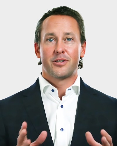 A man in a dark suit and white shirt gestures with his hands while speaking, against a plain background.