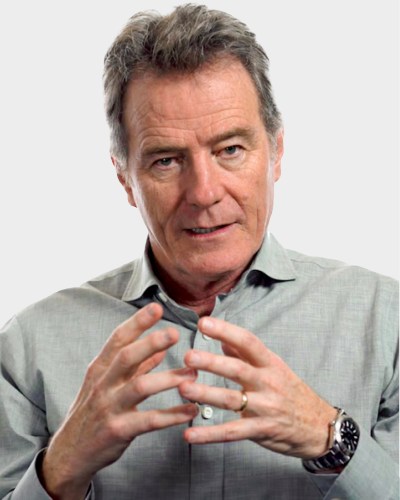 A man with gray hair and a serious expression gestures with his hands while wearing a gray shirt and a wristwatch against a plain background.