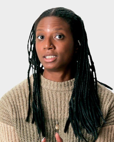 A person with long braided hair and wearing a knit sweater and necklace, speaks in front of a plain background.