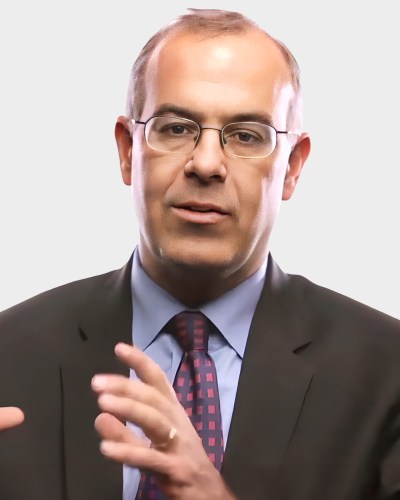 A man with glasses, wearing a suit and tie, gestures with both hands while talking against a plain background.