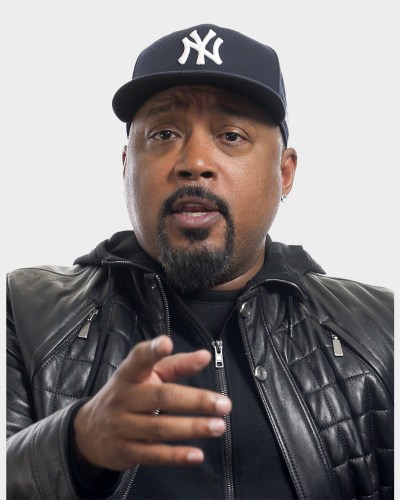 A man wearing a New York Yankees cap, a leather jacket, and a black shirt gestures while looking ahead.