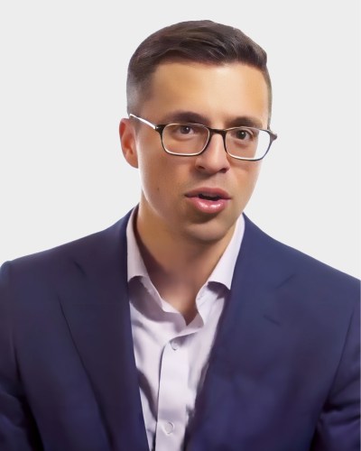 A man with short brown hair, wearing glasses, a white dress shirt, and a dark suit jacket.