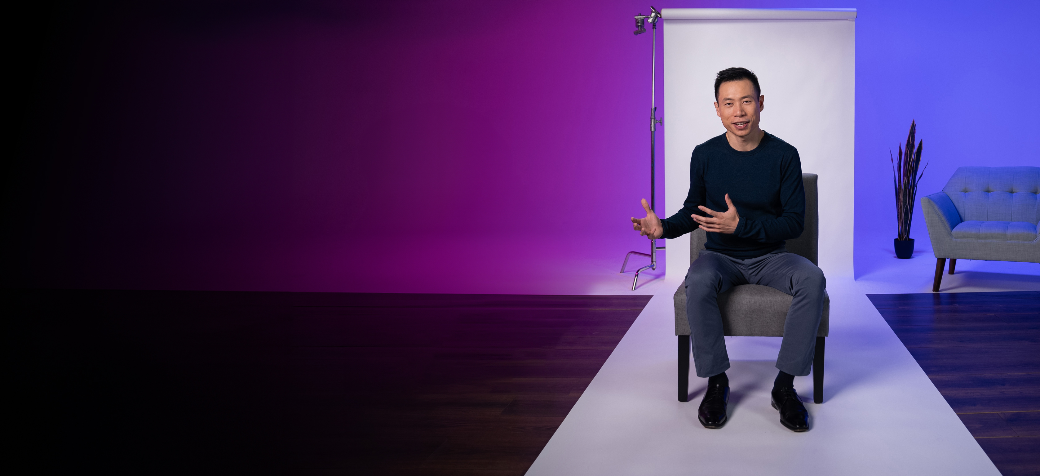 A person sits on a chair in a studio with a purple and blue lighting background, gesturing with their hands. A gray sofa and a plant are visible in the background.