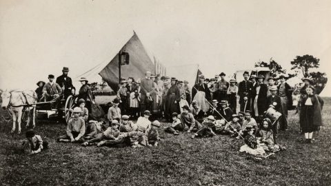 A large group of people, some standing and some sitting, gather outdoors in front of a tent and a horse-drawn carriage, set against a grassy field with a few trees in the background.