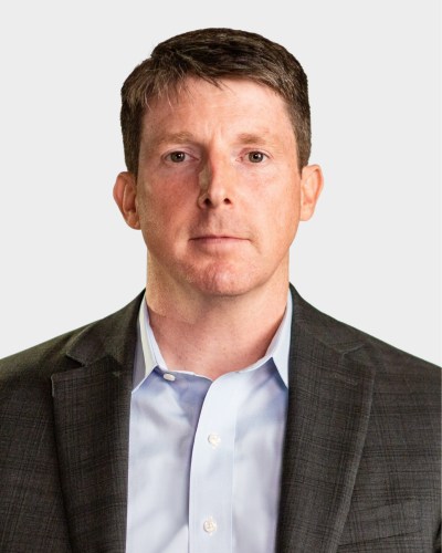 A man with short brown hair in a dark suit and light blue shirt looks directly at the camera against a plain background.