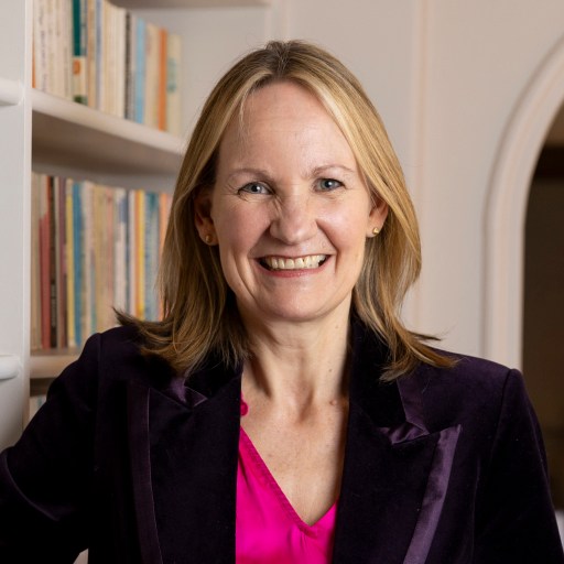 A woman with blonde hair and a bright smile is standing in front of a bookshelf. She is wearing a dark blazer and a pink top.