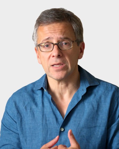 A man with glasses and gray hair is talking. He is wearing a blue shirt and gesturing with his hands. The background is plain and light-colored.