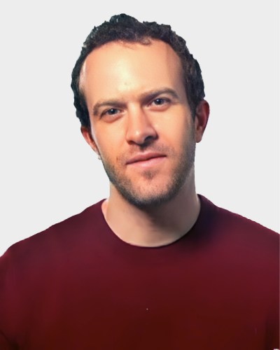 A man with short curly hair and a trimmed beard, wearing a red shirt, stands against a plain white background.