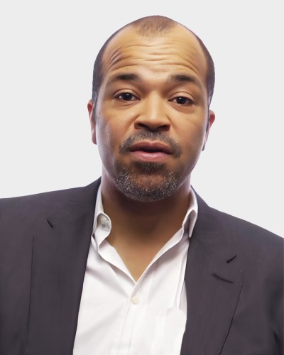 A portrait of a man with a closely shaved head and trimmed beard, wearing a black suit jacket and a white shirt, posing against a plain white background.