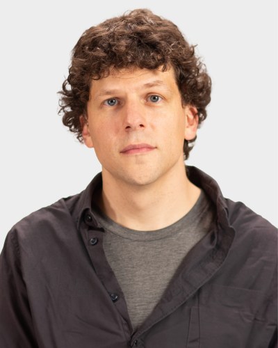 A person with curly brown hair wearing a black shirt over a gray t-shirt stands in front of a plain white background.