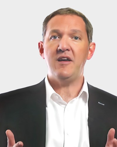 A man with short brown hair is wearing a black suit jacket and white dress shirt. He appears to be speaking, gesturing with both hands. The background is plain white.