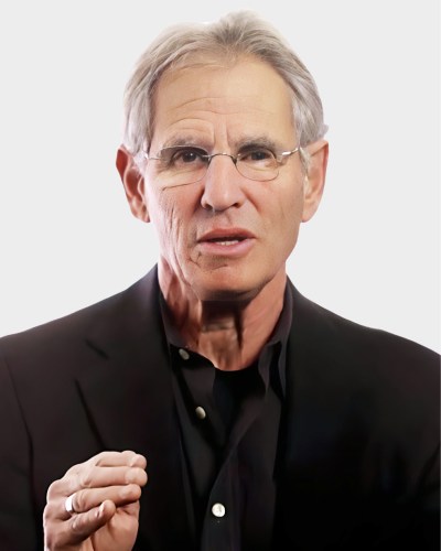 An older man with gray hair and glasses, wearing a black suit, gestures with his right hand while speaking against a plain white background.