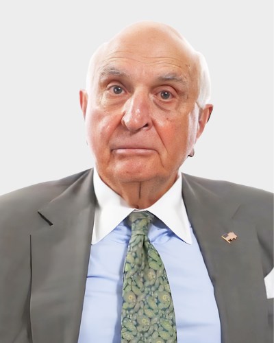 Bald elderly man in a suit and tie with a neutral expression, standing against a plain white background.