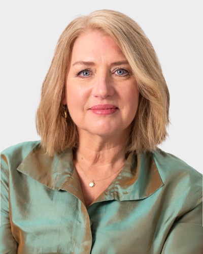 A middle-aged woman with shoulder-length blonde hair, wearing a teal button-up shirt, poses for a portrait against a plain white background.