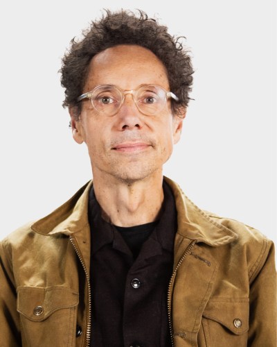 A person with short curly hair and glasses is wearing a brown jacket over a black shirt against a plain background.