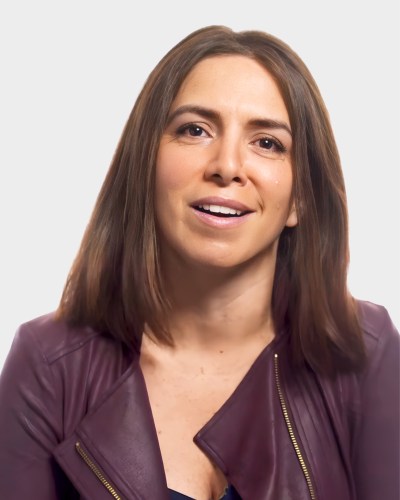 A person with shoulder-length hair, wearing a maroon jacket, is smiling in front of a plain white background.