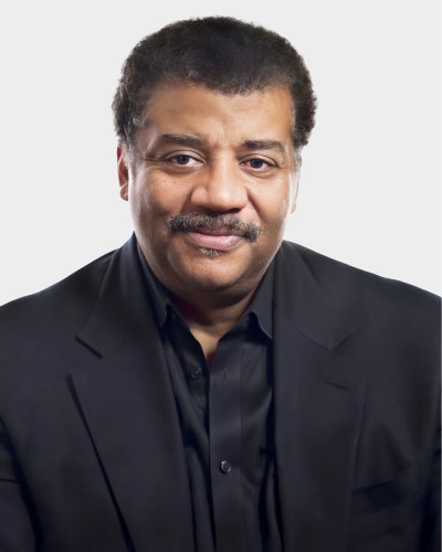 A man with short hair and a mustache, wearing a black shirt and black blazer, looks at the camera with a slight smile.