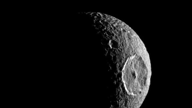 Image of Mimas, one of Saturn's moons, partially illuminated. The large Herschel crater is prominently visible on the right side of the moon's surface.