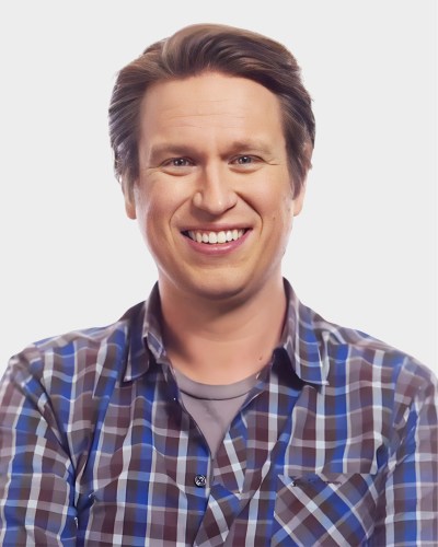 A person with short brown hair wearing a plaid shirt smiles at the camera against a plain background.