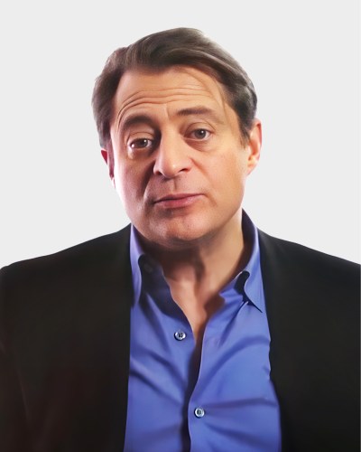 A person with short brown hair wearing a blue shirt and black jacket looks at the camera against a plain background.