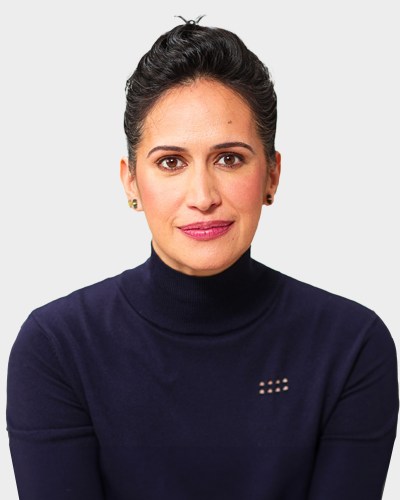 A person with dark hair in a bun, wearing a navy blue turtleneck, looking at the camera against a plain white background.