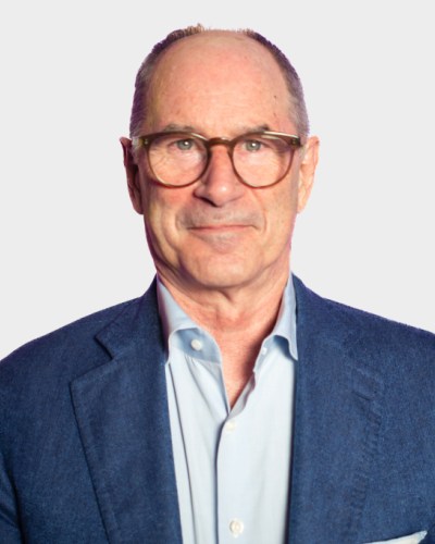 A man with glasses, wearing a blue jacket over a light blue shirt, stands against a plain background.