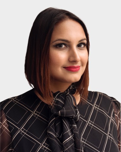 A person with shoulder-length hair, wearing a patterned black blouse with a neck tie, looks towards the camera with a neutral expression.