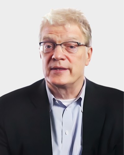 A middle-aged man with short, light-colored hair, wearing glasses, a light blue shirt, and a dark blazer is facing forward. The background is plain white.