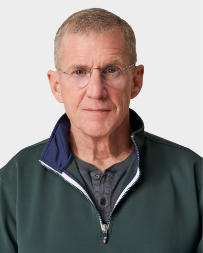 A middle-aged man with short gray hair and glasses is wearing a green zip-up collar jacket over a gray shirt, looking directly at the camera against a plain white background.