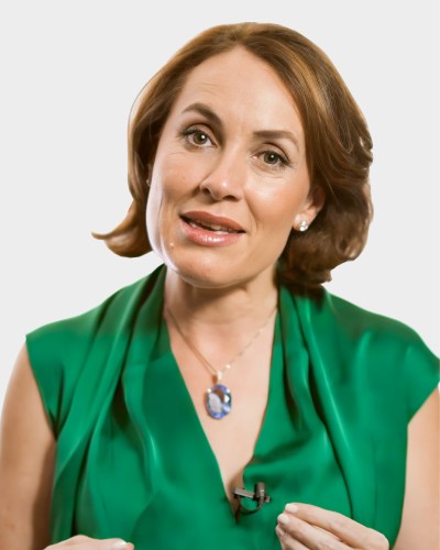 A woman wearing a green top and a pendant necklace speaks while gesturing with her hands. She has short brown hair styled in waves.