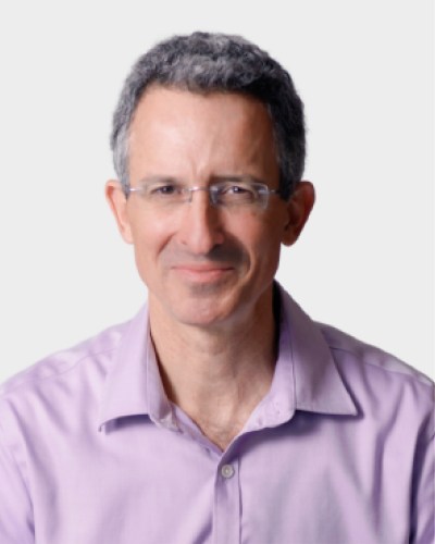 A man with short gray hair, wearing glasses and a light purple collared shirt, smiles softly while looking at the camera.