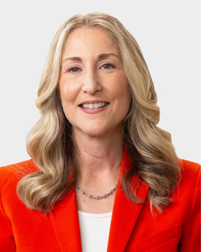 A woman with long, wavy blonde hair is smiling. She is wearing an orange blazer over a white top and a delicate necklace. The background is plain white.