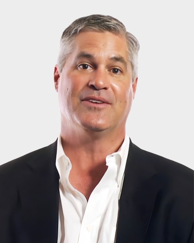 A man with short gray hair wearing a white shirt and dark suit jacket, facing the camera against a plain background.