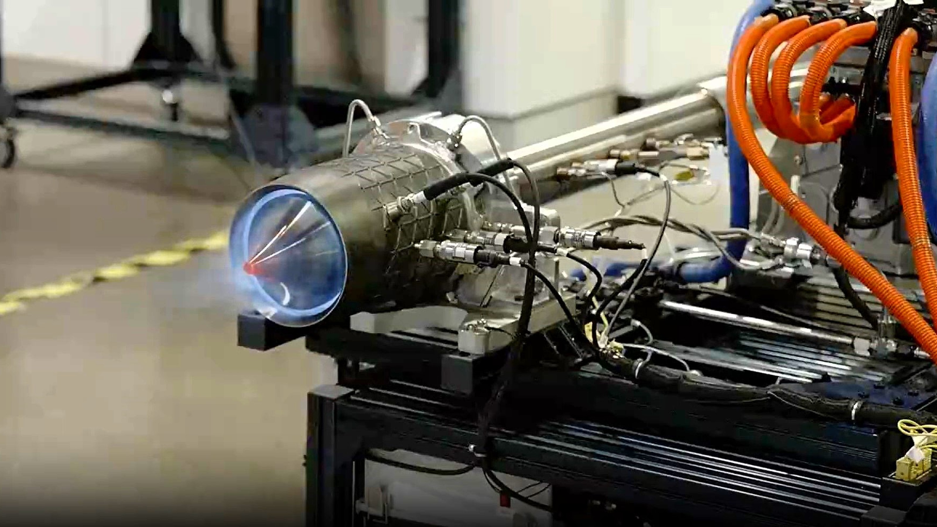 A close-up of a rocket engine testing setup, showing a small blue flame at the nozzle and multiple cables and pipes connected to the engine.