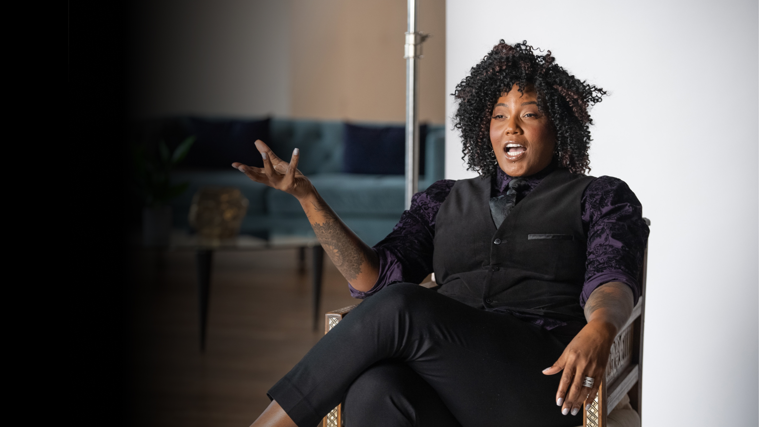 Person with curly hair sits in a chair, gesturing with one hand while speaking. The background includes a partially visible room with a sofa and a potted plant.