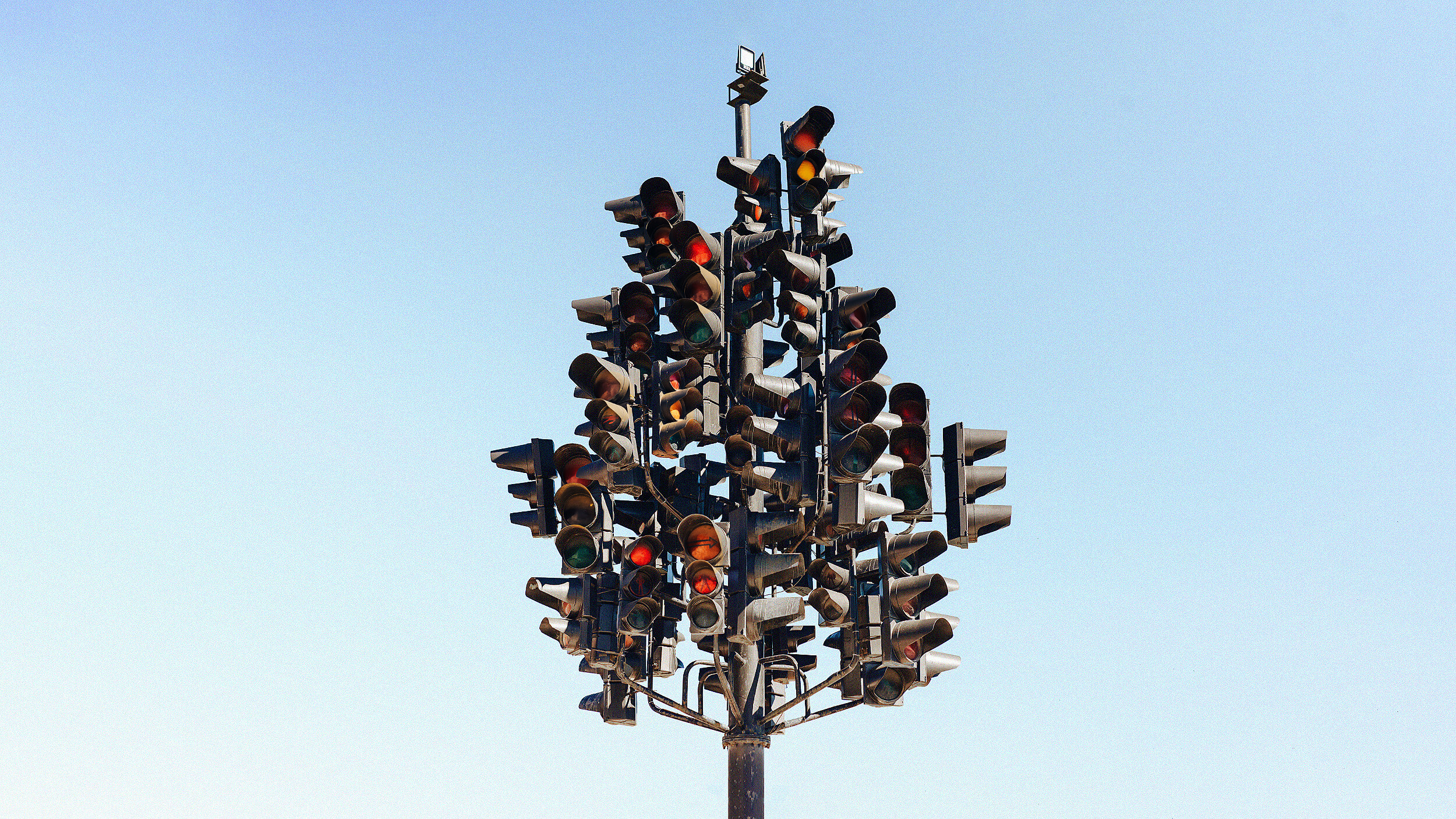 A pole with numerous traffic lights mounted in different directions creates an illusion of control against a clear sky background.