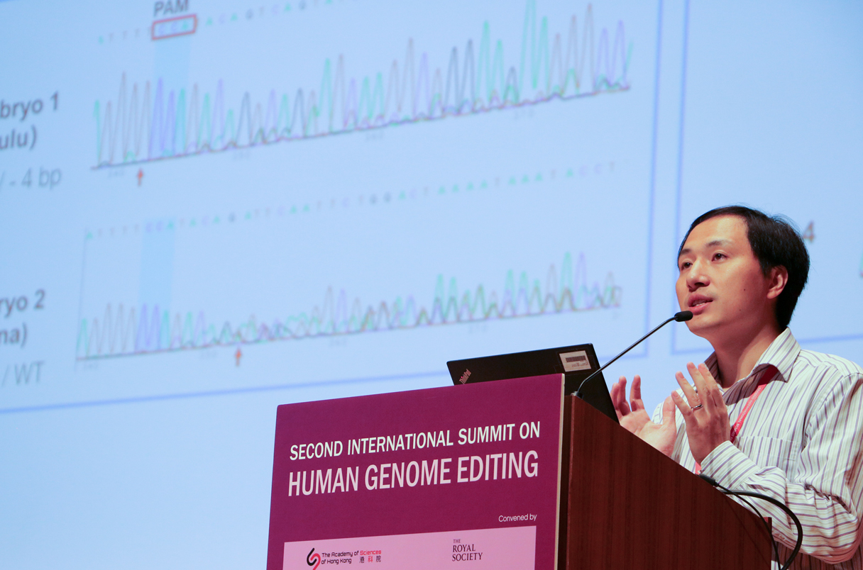 A speaker presents on human genome editing at the Second International Summit, with a scientific chart displayed on the screen behind.