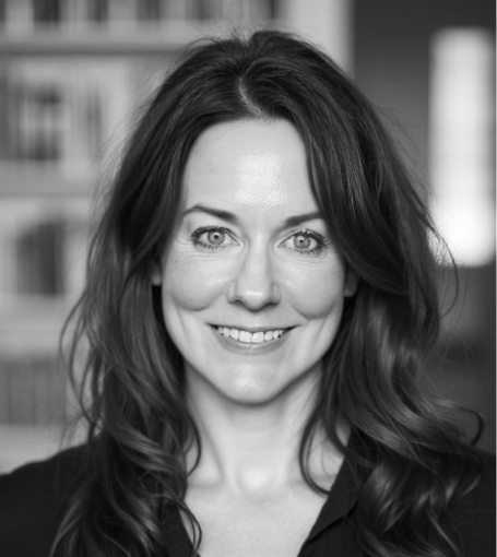 A woman with long hair smiles at the camera against a blurred background of a bookshelf. The image is in black and white.