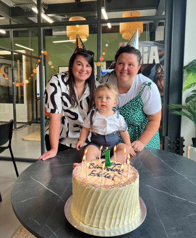 Two adults in party hats stand behind a toddler seated on a chair in front of a birthday cake with lit candles. The cake has the text "Happy Birthday" and the number one on it.