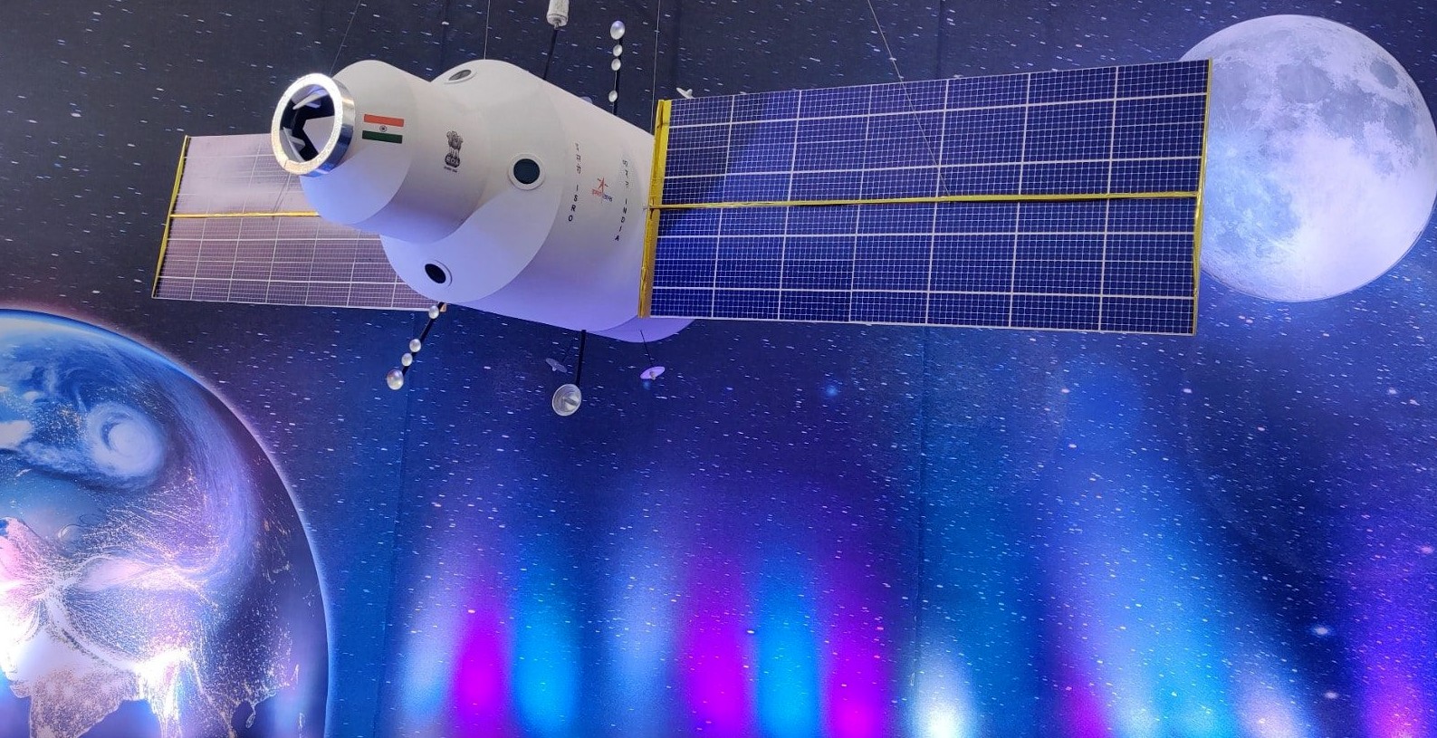 A model of a satellite in space with solar panels extended, the Earth and a bright moon visible in the background. The satellite has an Indian flag and markings on its body.